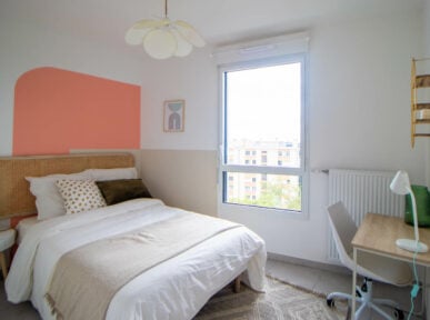 Chambre à louer à Lyon (69) par Colivys, mur orange, lit avec cadre en bois, fenêtre, bureau avec lampe de bureau, chaise blanche, tapis à formes géométriques